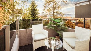 Un balcón con sillas de mimbre y una mesa de vidrio, con vistas a una zona residencial con árboles y edificios.