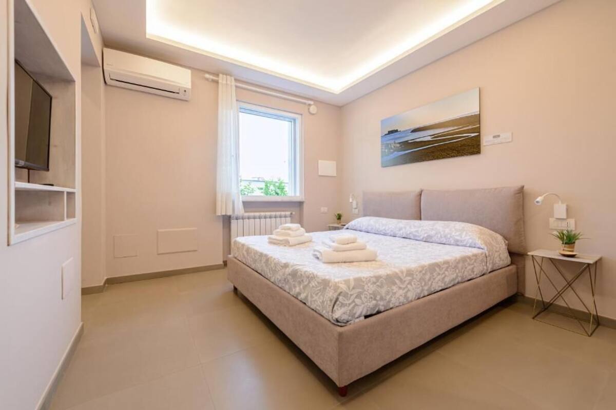 Egy modern hálószoba, amelyben található egy ágy, egy ablak, egy légkondicionáló, egy festmény, egy éjjeliszemélyke lámpával és egy kis növény.