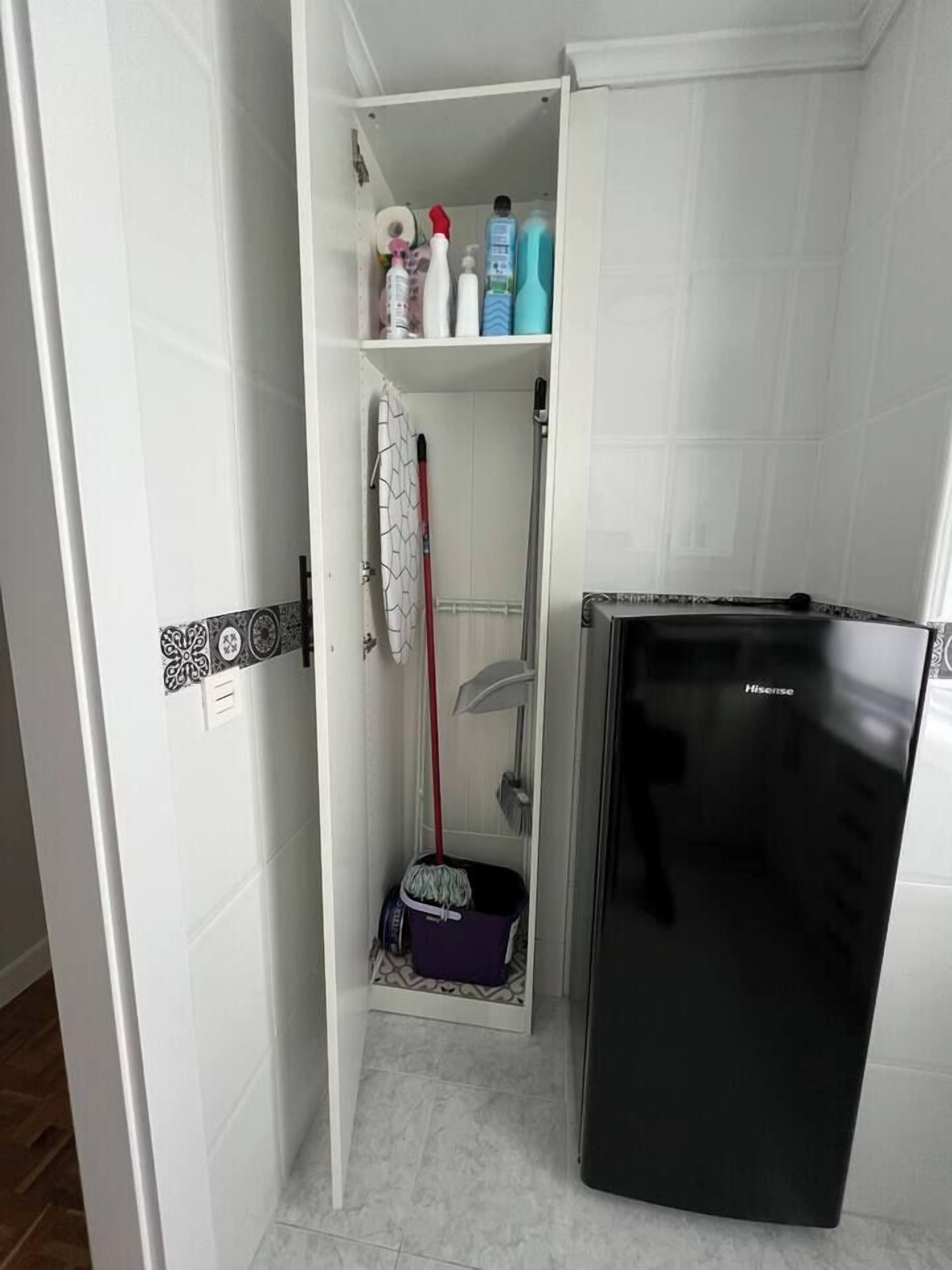 Une petite salle de bain avec un réfrigérateur noir, une serpillière et des produits de nettoyage rangés sur les étagères.