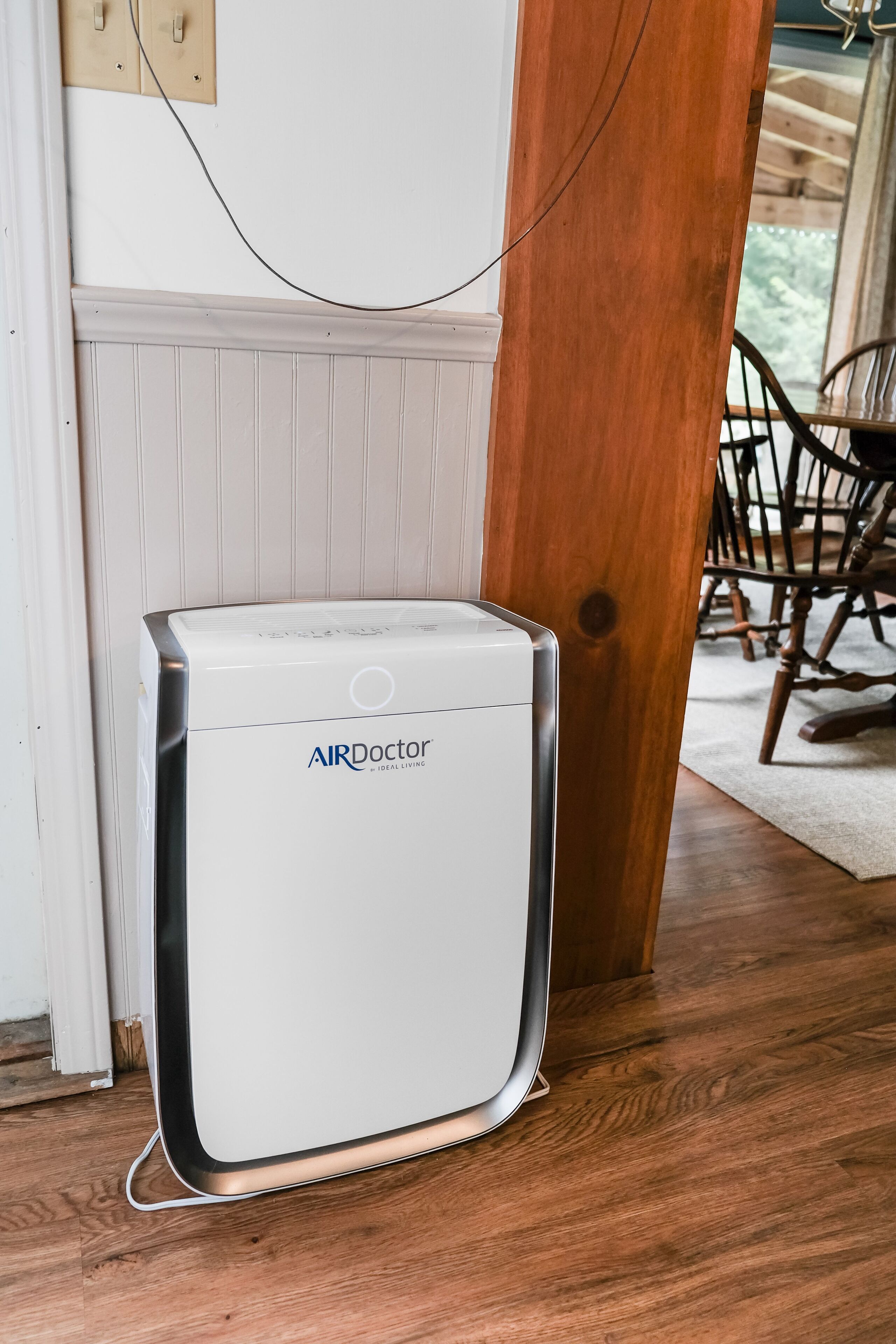 Un purificatore d'aria bianco con il marchio 'AIRDoctor' in una stanza con pavimento in legno e una zona pranzo visibile sullo sfondo.