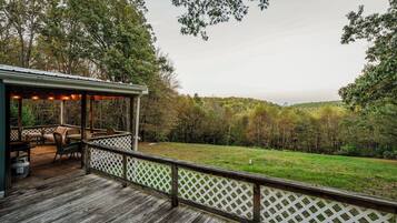 A wooden deck with outdoor furniture and a covered seating area overlooking a grassy field and forested hills.