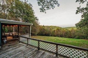 Eine Holzterrasse mit Gartenmöbeln und einem überdachten Sitzbereich mit Blick auf eine Wiese und bewaldete Hügel.
