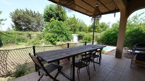 Une terrasse extérieure avec une table et des chaises en bois, entourée d’une clôture et d’une végétation luxuriante.