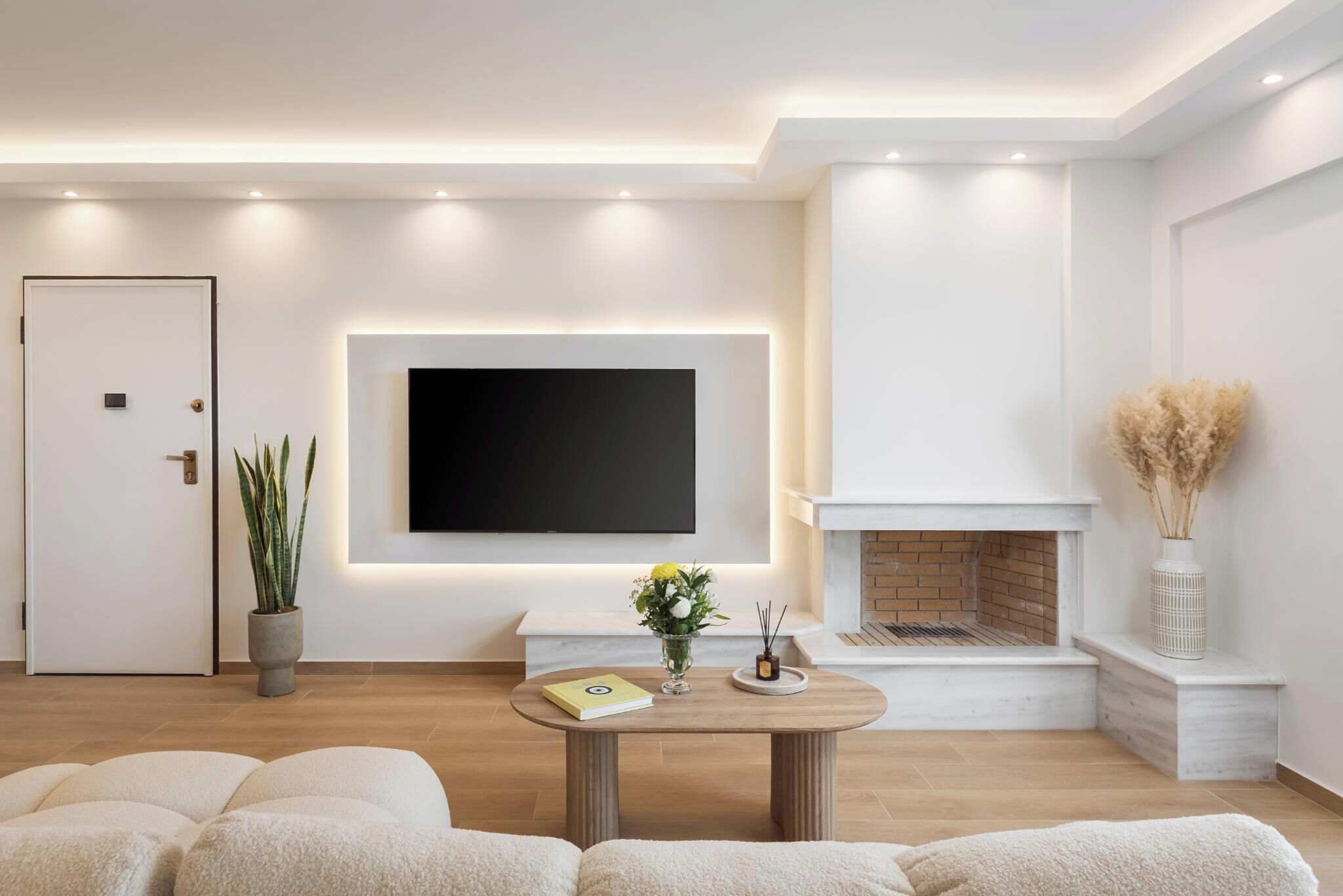 Ein modernes Wohnzimmer mit einem an der Wand montierten Flachbildfernseher, einem Kamin und einem runden Couchtisch mit Buch und Blumen.