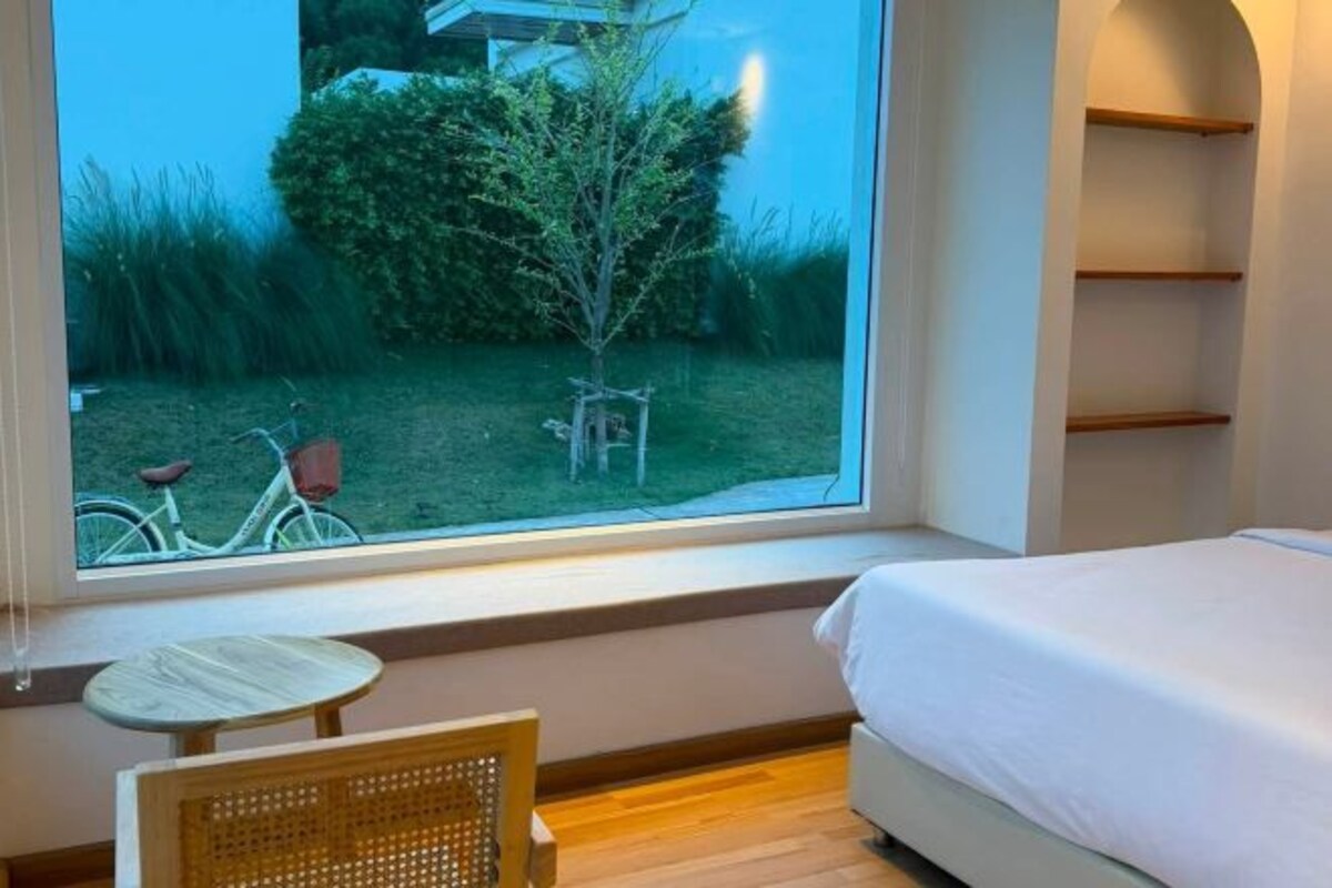 A bedroom with a bed, a window with a view of a garden, a small table, and a chair.