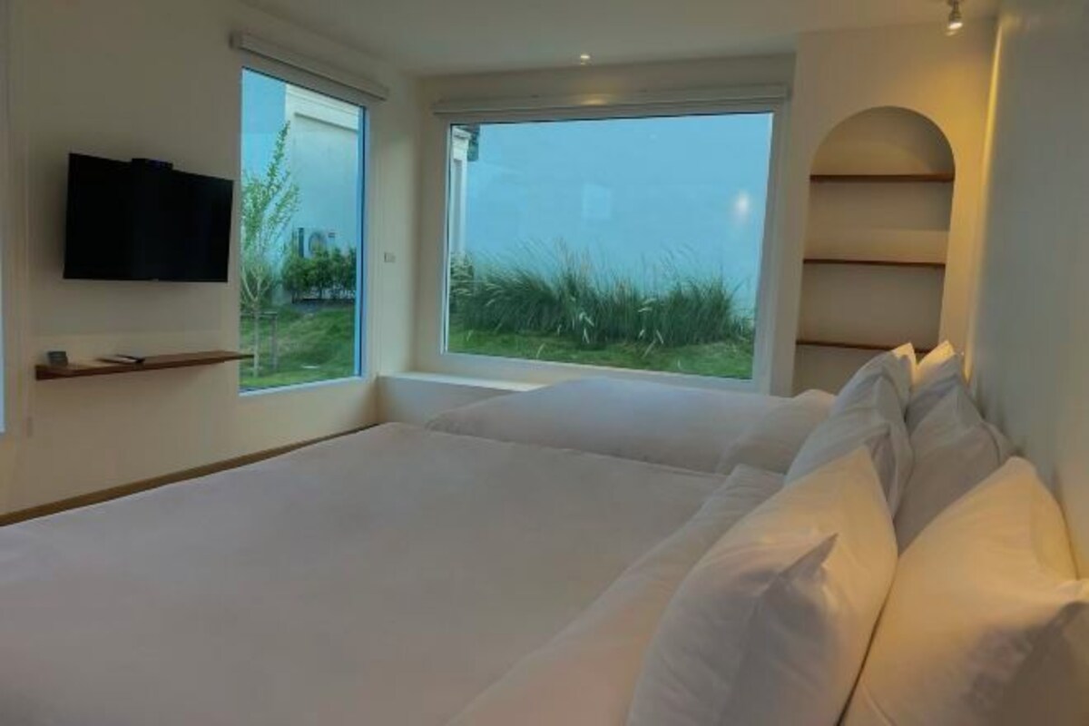 A bedroom with a large bed, a TV mounted on the wall, and a window with a view of grass and trees.