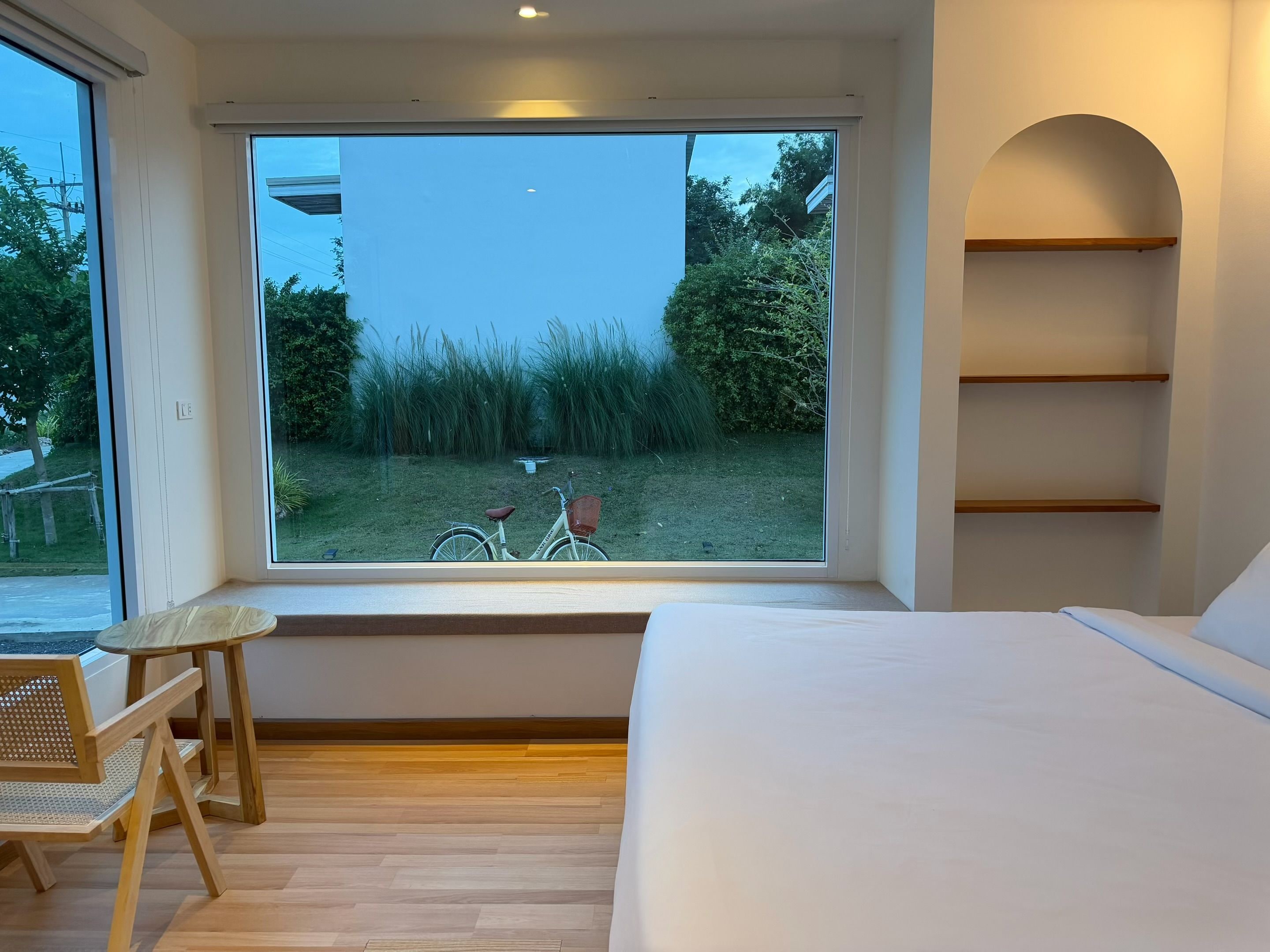 A bedroom with a large window, a bed, a wooden chair, and a bicycle outside.