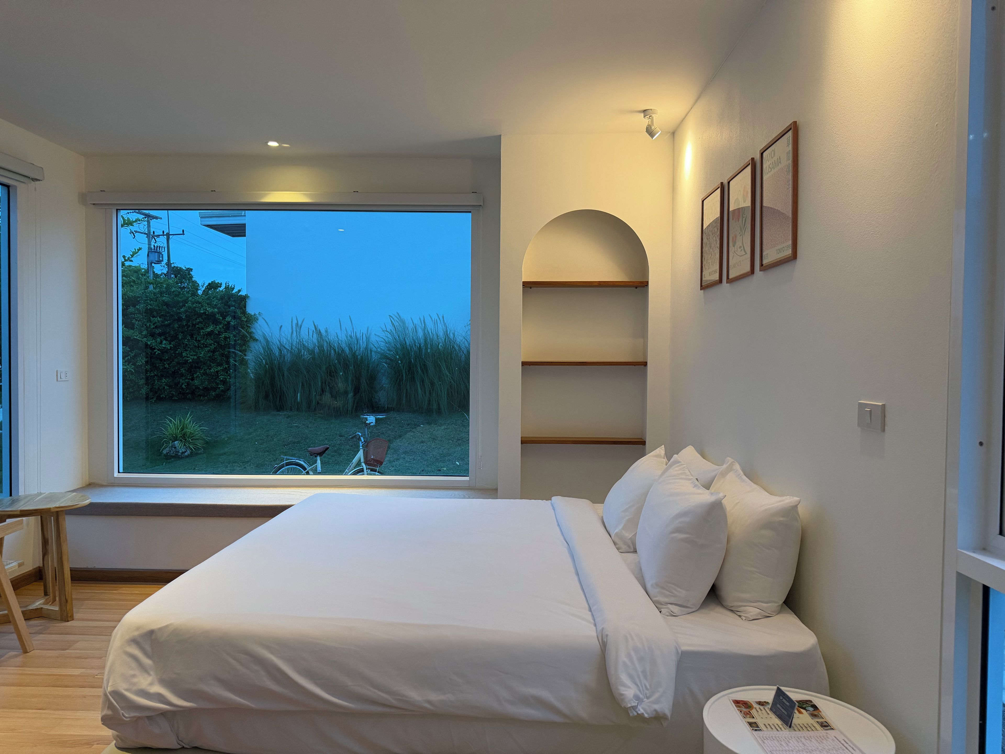 Un dormitorio con un ventanal grande, una cama con ropa de cama blanca, una mesita de noche y vistas al exterior con una bicicleta y vegetación.