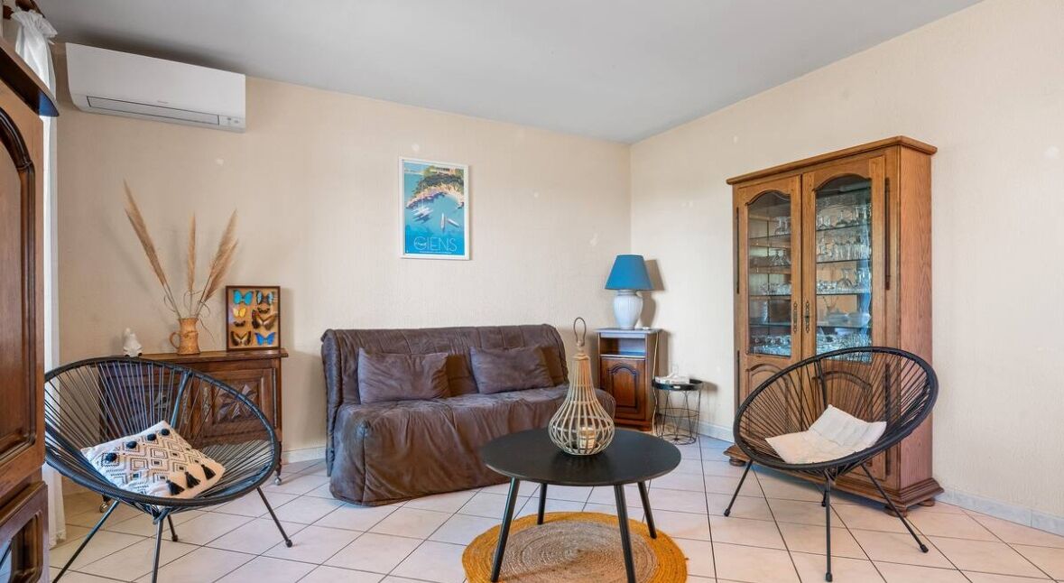 A living room with a brown sofa, two black wire chairs, a small round table, a cabinet with glass doors, and a wall-mounted air conditioner.