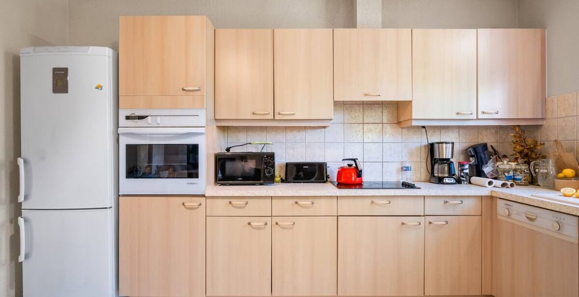 A kitchen with light wooden cabinets, a white refrigerator, a microwave, a kettle, and various kitchen appliances on the counter.