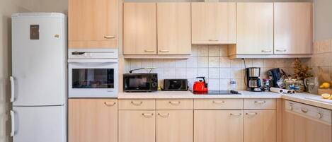 A kitchen with light wooden cabinets, a white refrigerator, a microwave, a kettle, and various kitchen appliances on the counter.