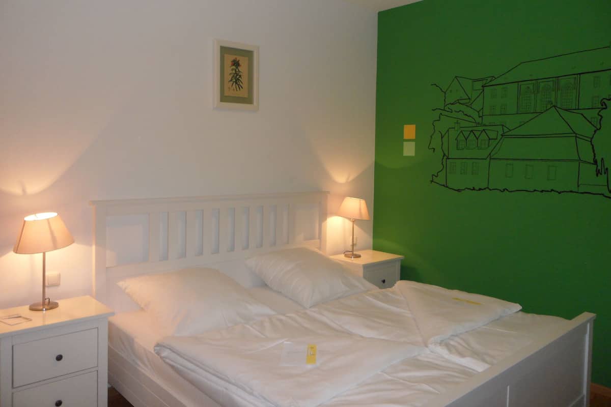 A neatly made bed with white linens, two bedside lamps, a small bedside table, and a green wall with a simple line drawing.