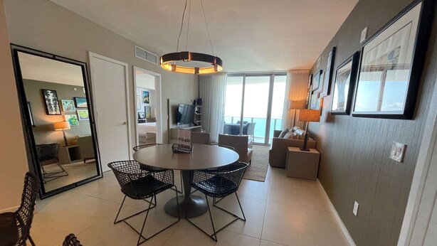 A modern hotel room with a round dining table, black wireframe chairs, a ceiling light fixture, a large mirror, and a view of the balcony.