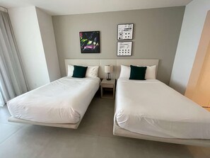 Habitación de hotel con dos camas individuales, una mesita de noche con lámpara, y una pared con arte abstracto y un enchufe.