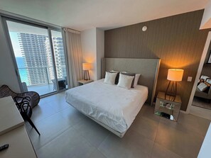 Et moderne hotelværelse med en stor seng, to sengeborde med lamper, en stol og udsigt til bybilledet gennem et stort vindue.