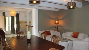 Un comedor con una mesa de madera, sillas y una mesa auxiliar de vidrio. Se observa un sofá y una lámpara al fondo.