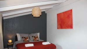 A bedroom with a bed, pillows, a red blanket, a hanging light fixture, and a red painting on the wall.