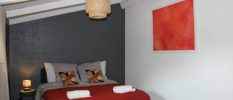 Un dormitorio con una cama, almohadas, una colcha roja, una lámpara colgante y un cuadro rojo en la pared.