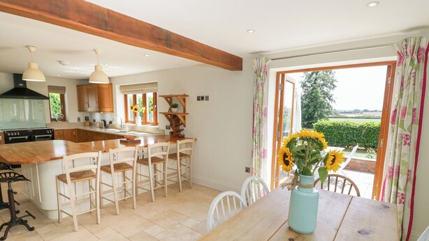 Une cuisine avec des meubles en bois, un îlot central doté de tabourets de bar, une table à manger ornée d’un vase de tournesols et une vue sur un jardin, visible à travers une grande fenêtre.