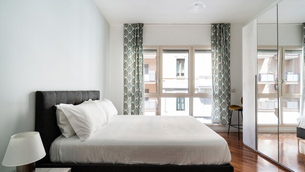 A modern bedroom with a large bed, a bedside table with a lamp, and a view of the outside through a sliding glass door. - Levante 65 by Wonderful Italy (Bari)
