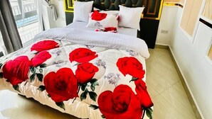 Ein Schlafzimmer mit einem Bett, grauen Vorhängen, einem schwarzen Kopfteil und einer blumigen Tagesdecke.