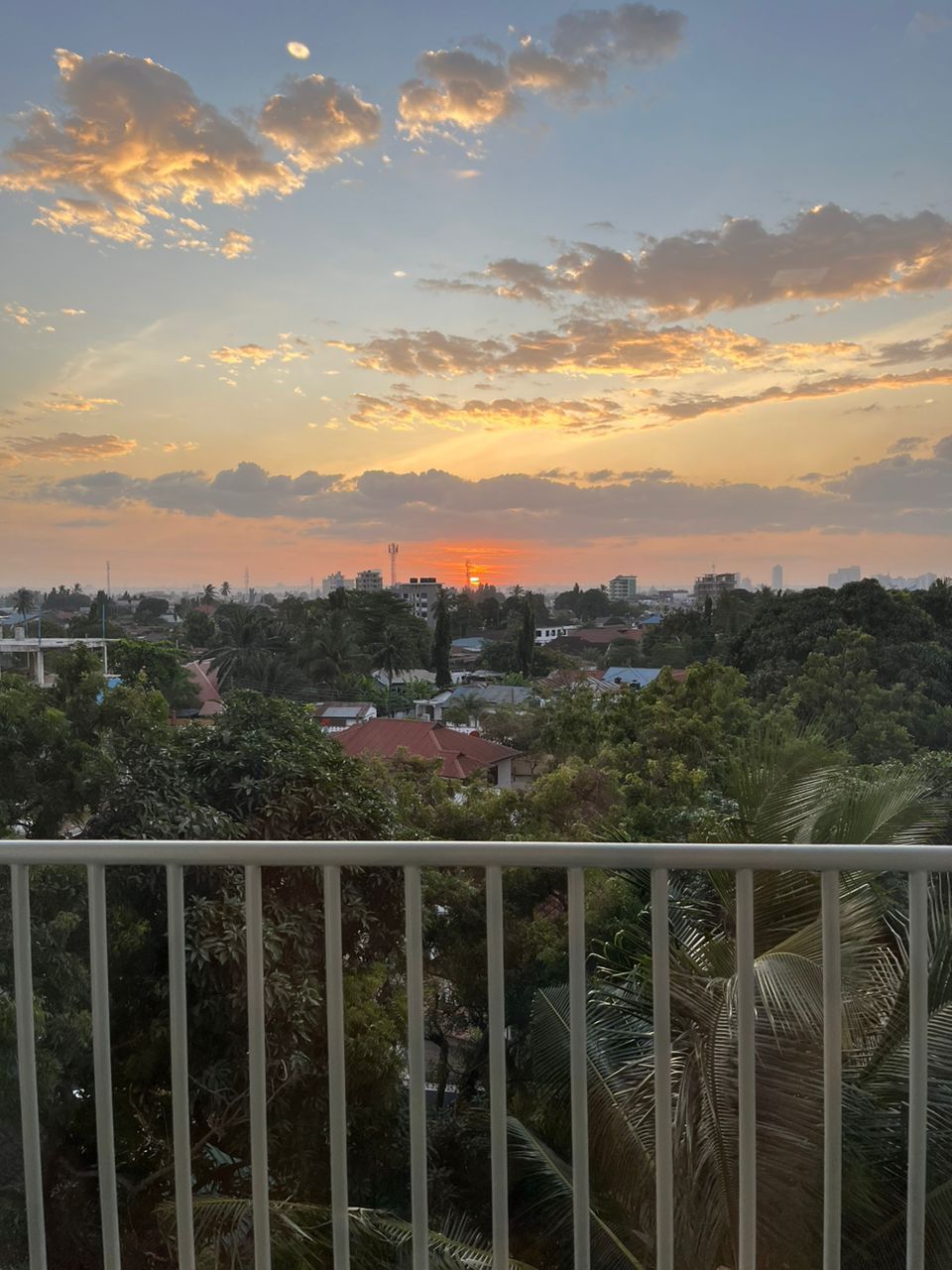 Une vue sur le coucher de soleil depuis un balcon doté d’une balustrade, donnant sur un quartier résidentiel avec des maisons et des arbres.