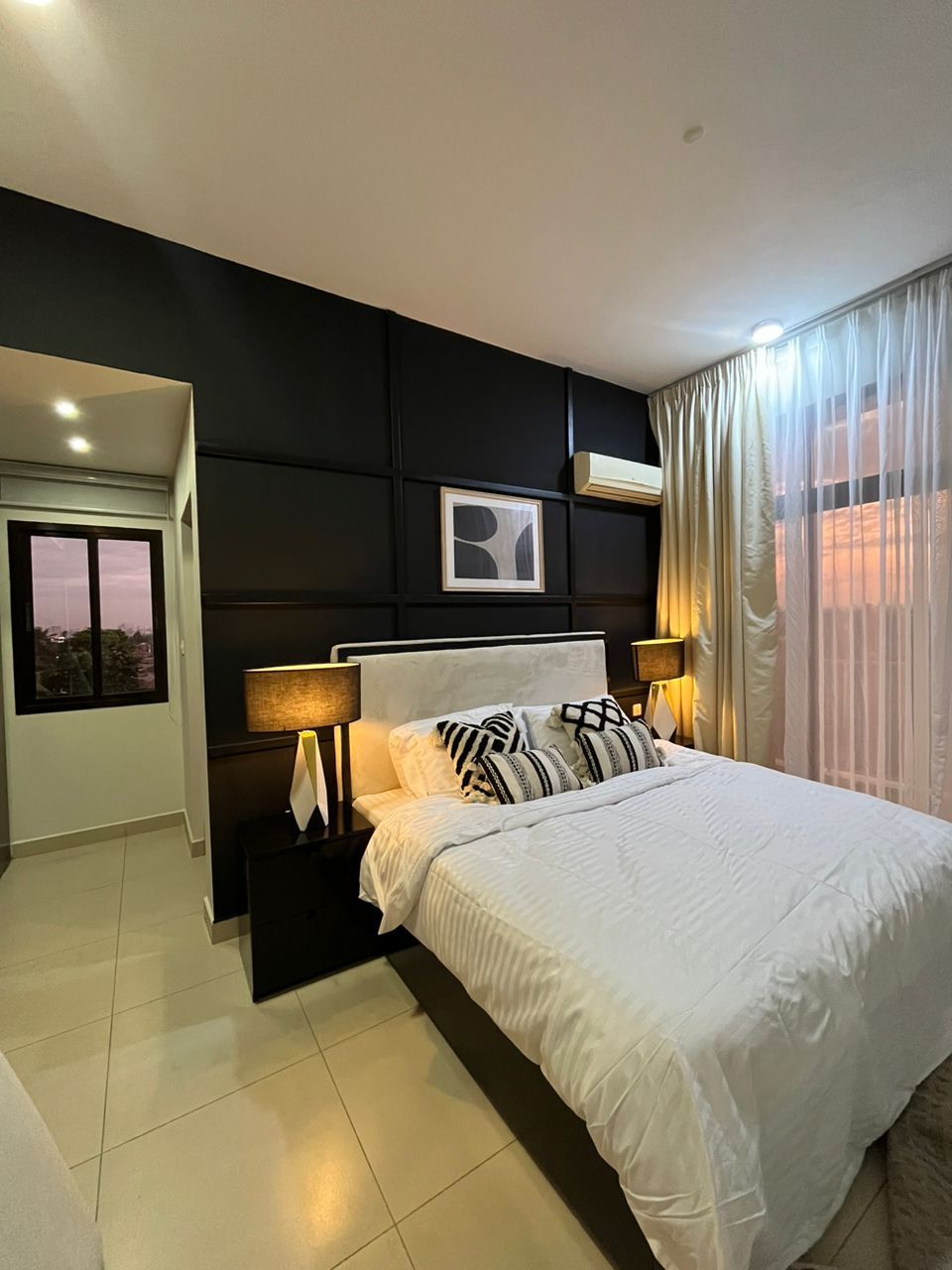 A modern hotel room with a large bed, two bedside tables with lamps, a black headboard, a framed picture, and a window with curtains.