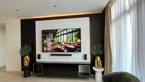 Ein modernes Wohnzimmer mit einem großen Flachbildfernseher an der Wand, einem dunkelgrünen Sofa, zwei Beistelltischen mit Zimmerpflanzen und einem gemusterten Teppich.