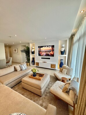 A modern living room with a large flat-screen TV, beige sofas, a coffee table, and decorative vases. - 3 bedroom apartment (Dar es Salaam)