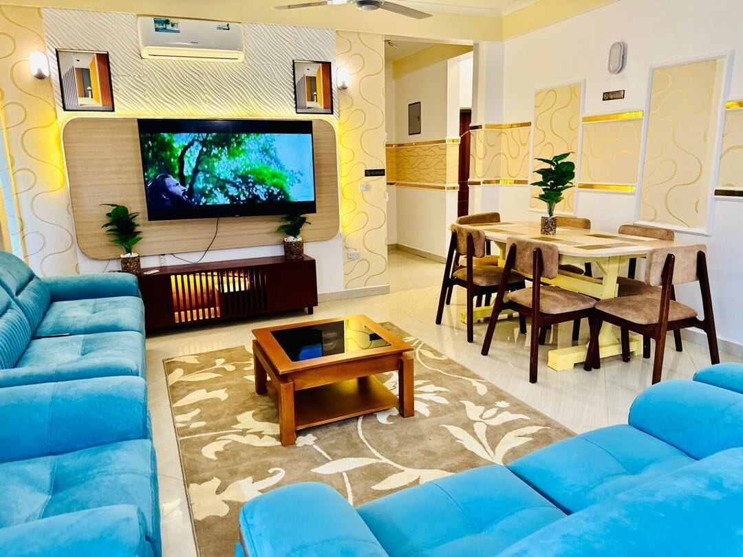 A modern living room with a flat-screen TV, blue sofas, a wooden coffee table, and a dining area with chairs and a table.