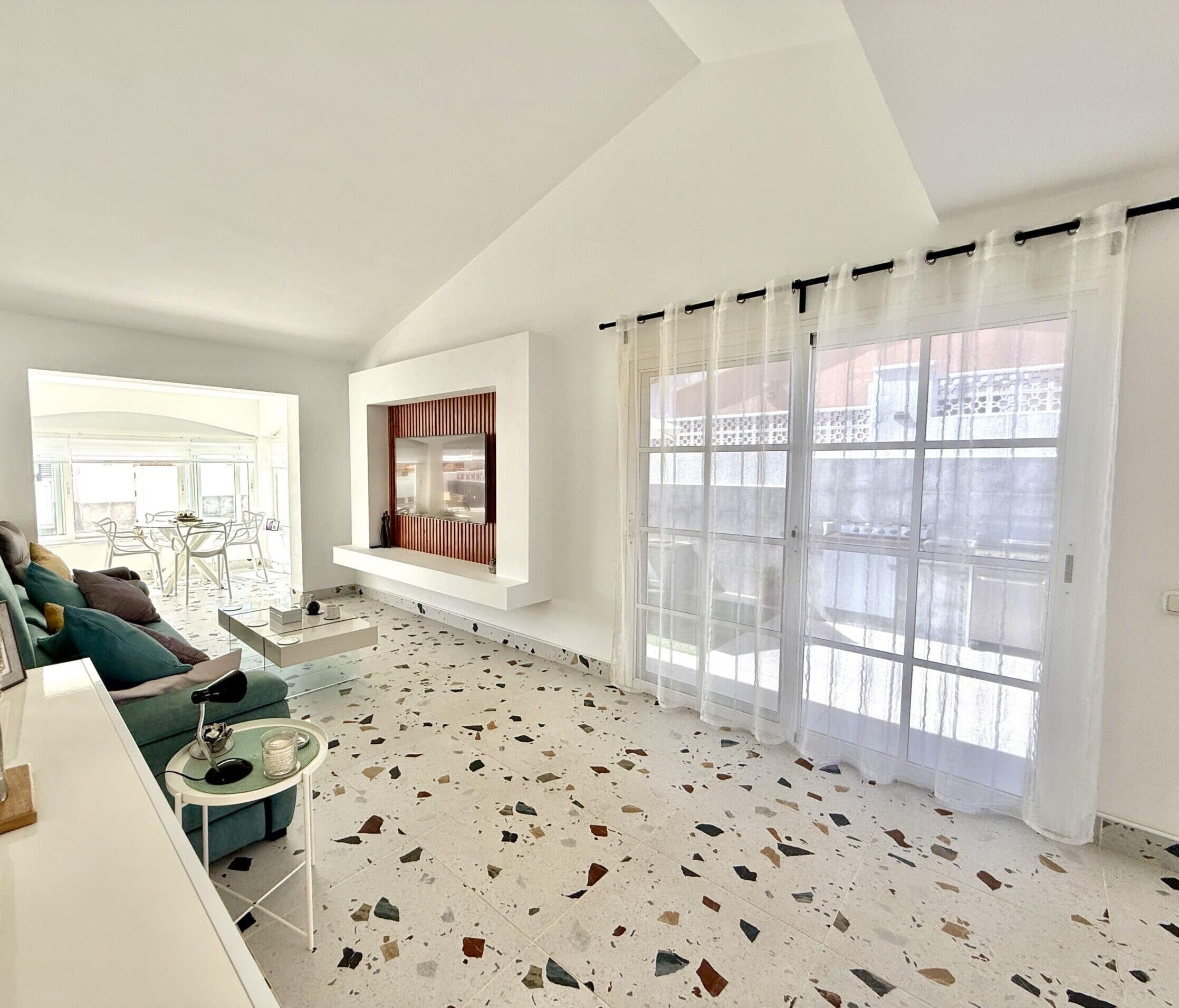 Een ruime woonkamer met een hoge plafond, een groot raam met gordijnen, een ingebouwde televisie en een terrazzovloer.