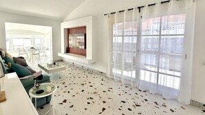 A spacious living room with a high ceiling, a large window with curtains, a built-in TV, and a terrazzo floor. - Villa Stay&Enjoy (Caleta de Fuste)