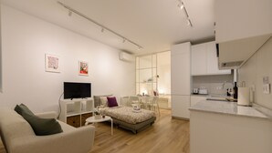 Ein modernes, helles Wohnzimmer mit einer Couch, einem kleinen Tisch und einem Fernseher. Es gibt eine Küchenzeile mit einer Arbeitsplatte und Schränken.