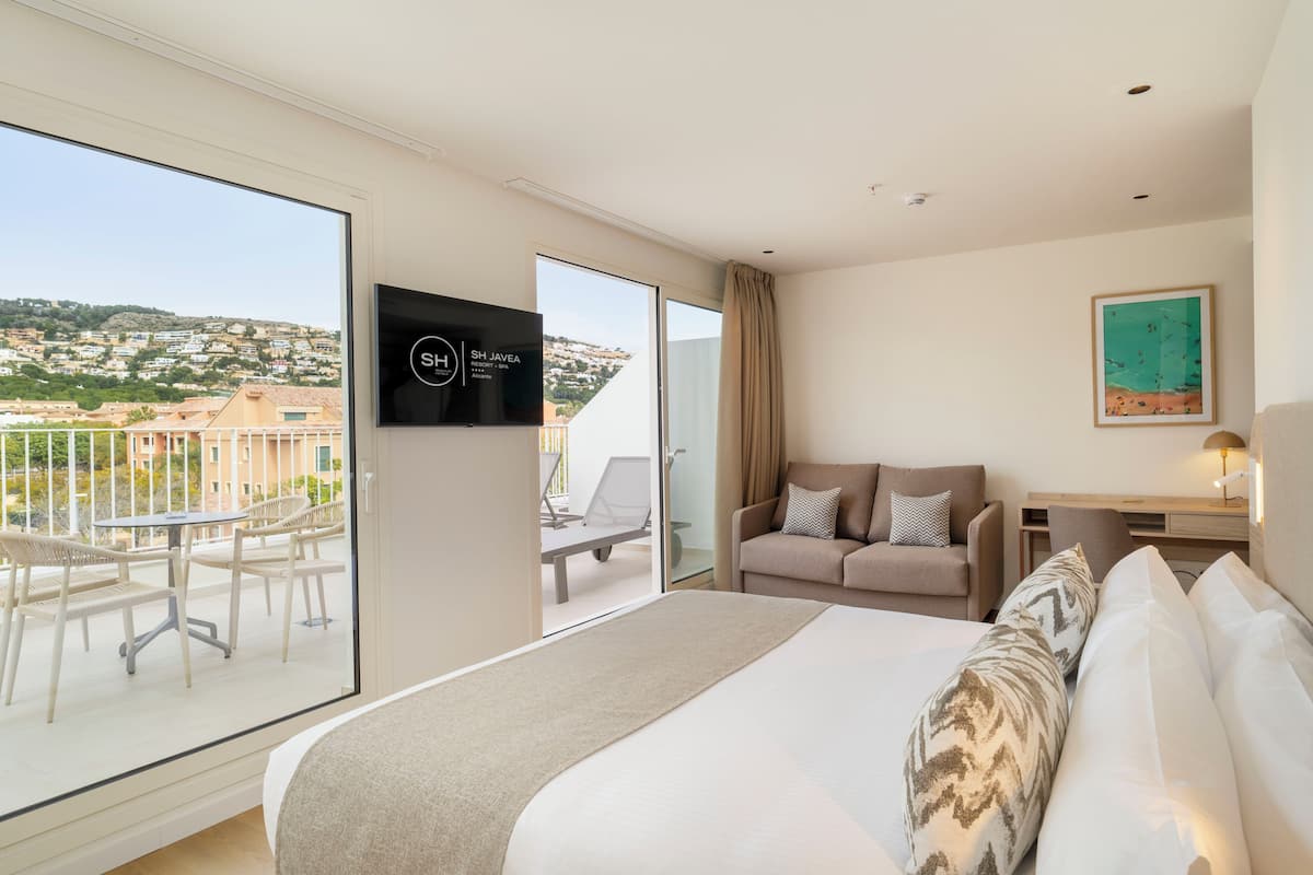 Ein Hotelzimmer mit Bett, Schreibtisch, Fernseher, Balkon mit Sitzgelegenheiten und Blick auf die Stadt.