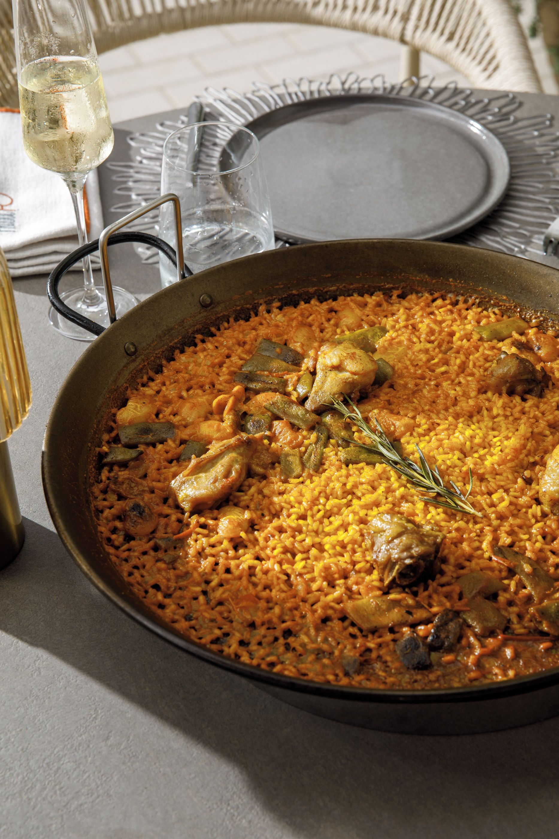 A large pan of paella with chicken and vegetables, a wine glass, and a dining table set with plates and cutlery.