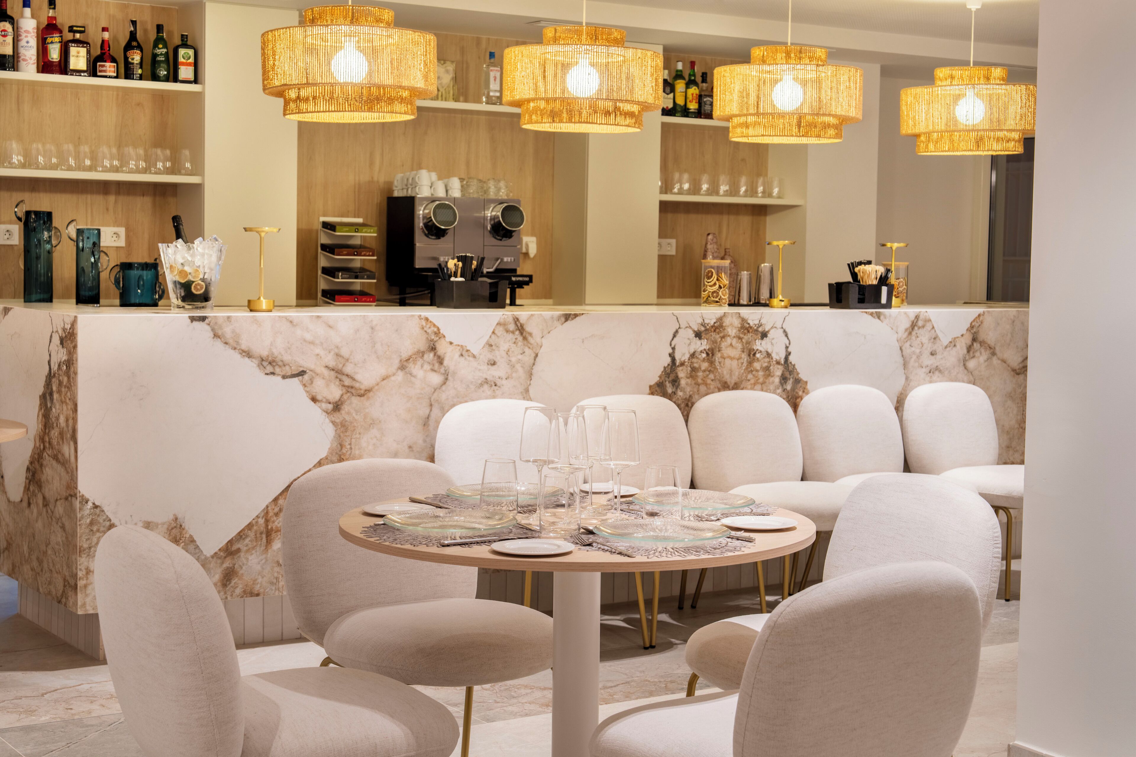 A bar area with a marble counter, wicker chandeliers, and seating with a marble wall backdrop.