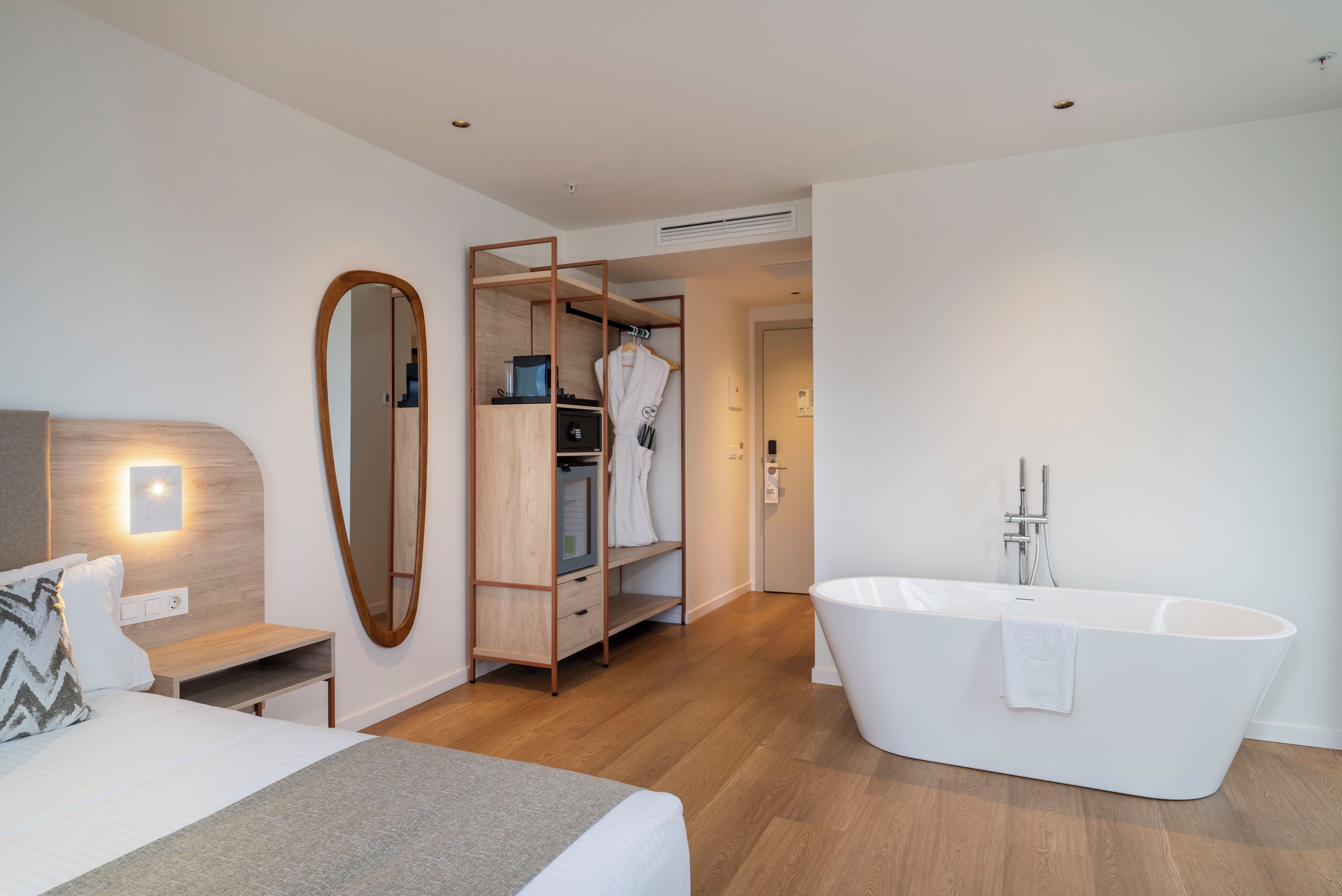 A modern hotel room with a large bed, a walk-in closet, a freestanding bathtub, and a mirror.