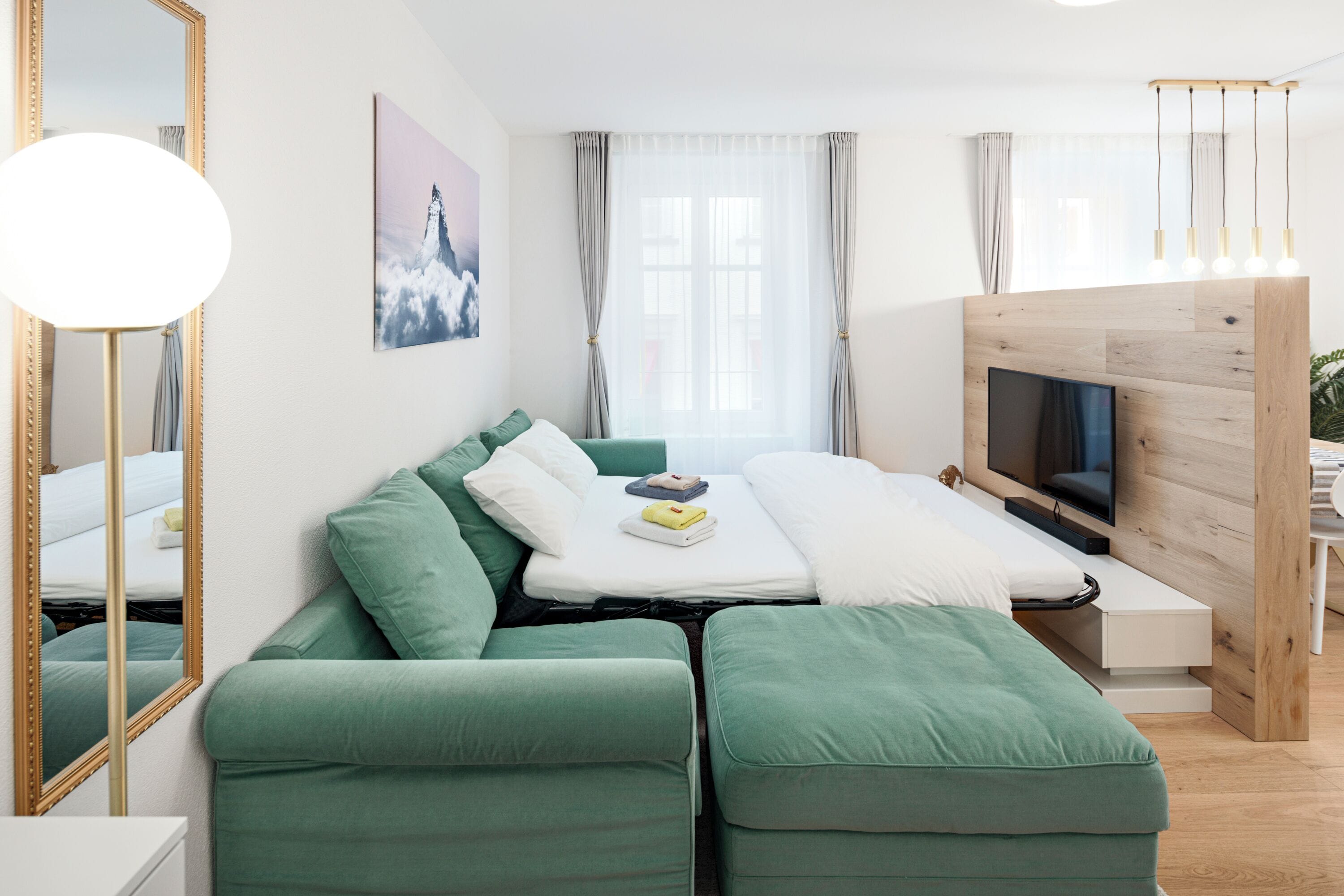 A modern hotel room with a green sofa, a flat-screen TV, a wooden TV stand, a large window with curtains, and a mirror with a gold frame.