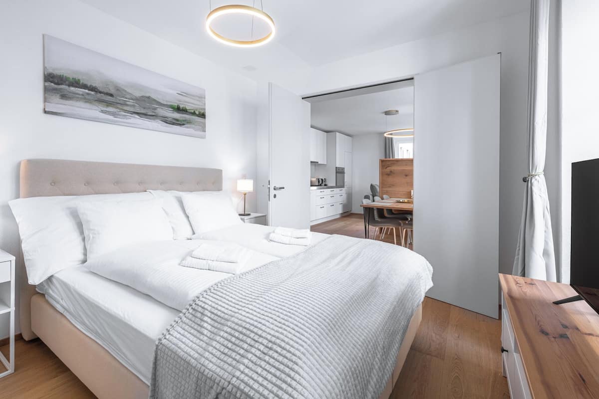 A modern bedroom with a large bed, a bedside table, a desk, and a view into the kitchen.