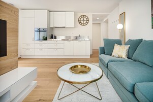 A modern living room with a teal sofa, a white coffee table, and a kitchen area in the background.