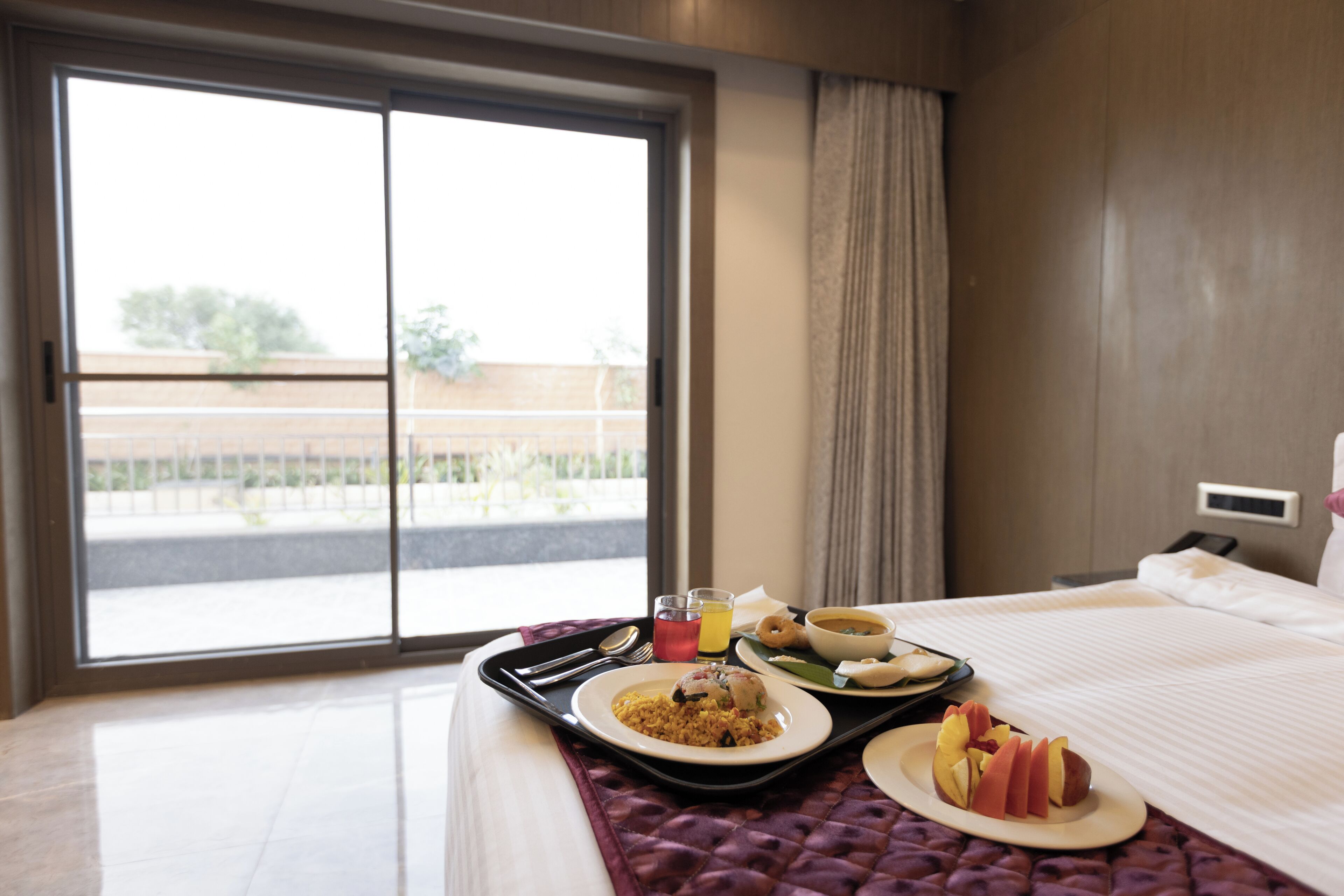 A hotel room with a large window, a bed with a white bedspread, a tray with breakfast items including eggs, toast, and fruit, and a view of a building and trees outside.