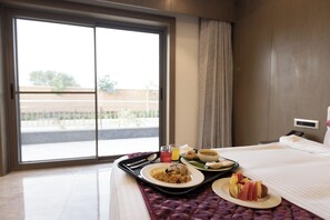A hotel room with a large window, a bed with a white bedspread, a tray with breakfast items including eggs, toast, and fruit, and a view of a building and trees outside. - Pride Resort Jodhpur (Jodhpur)