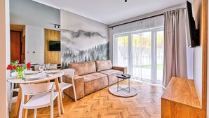 Una sala de estar moderna con suelo de madera en espiga, un sofá color beige y un comedor con sillas blancas y una mesa dispuesta para cuatro personas.