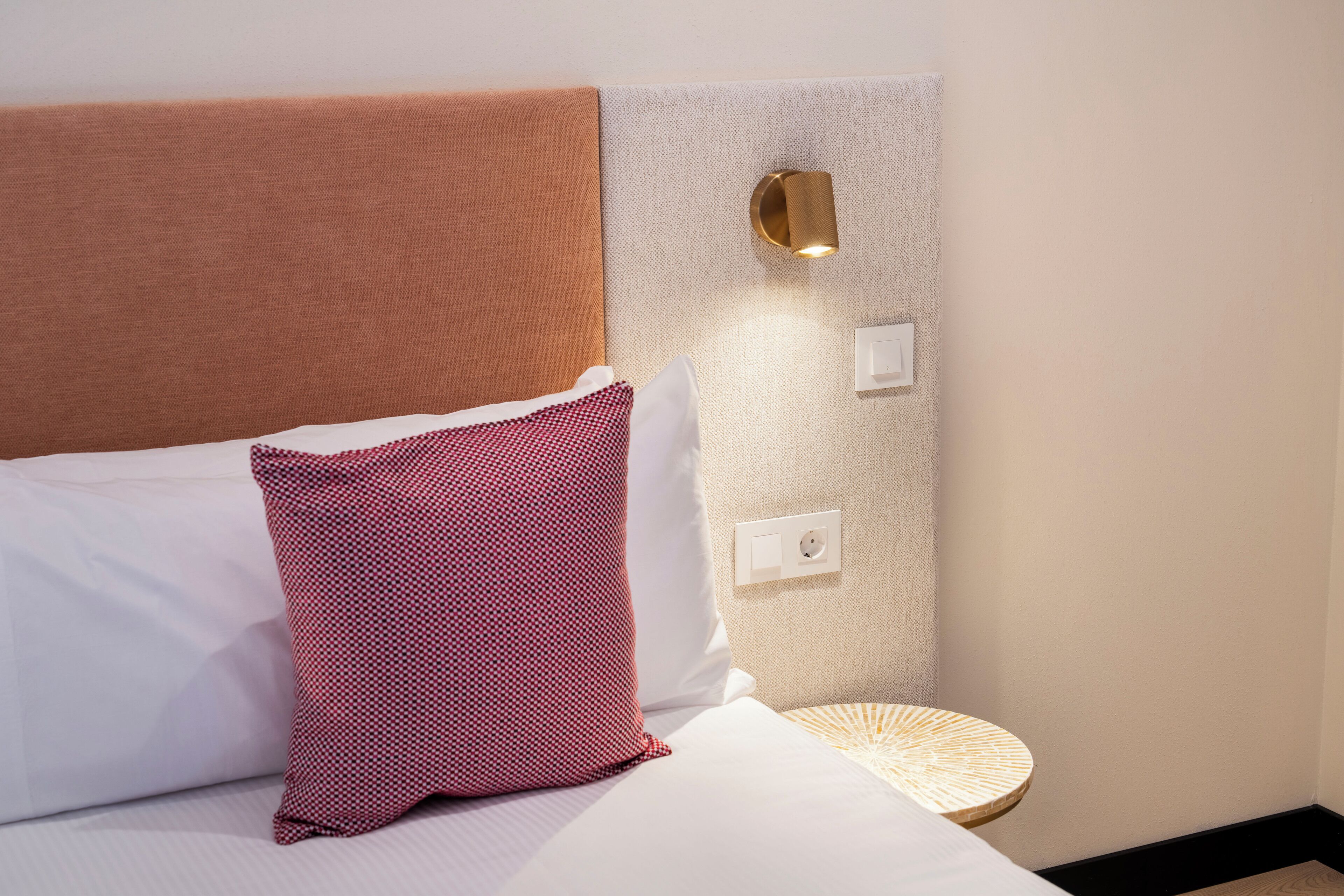 A neatly made bed with a patterned pillow, a round side table, and a wall-mounted light fixture.