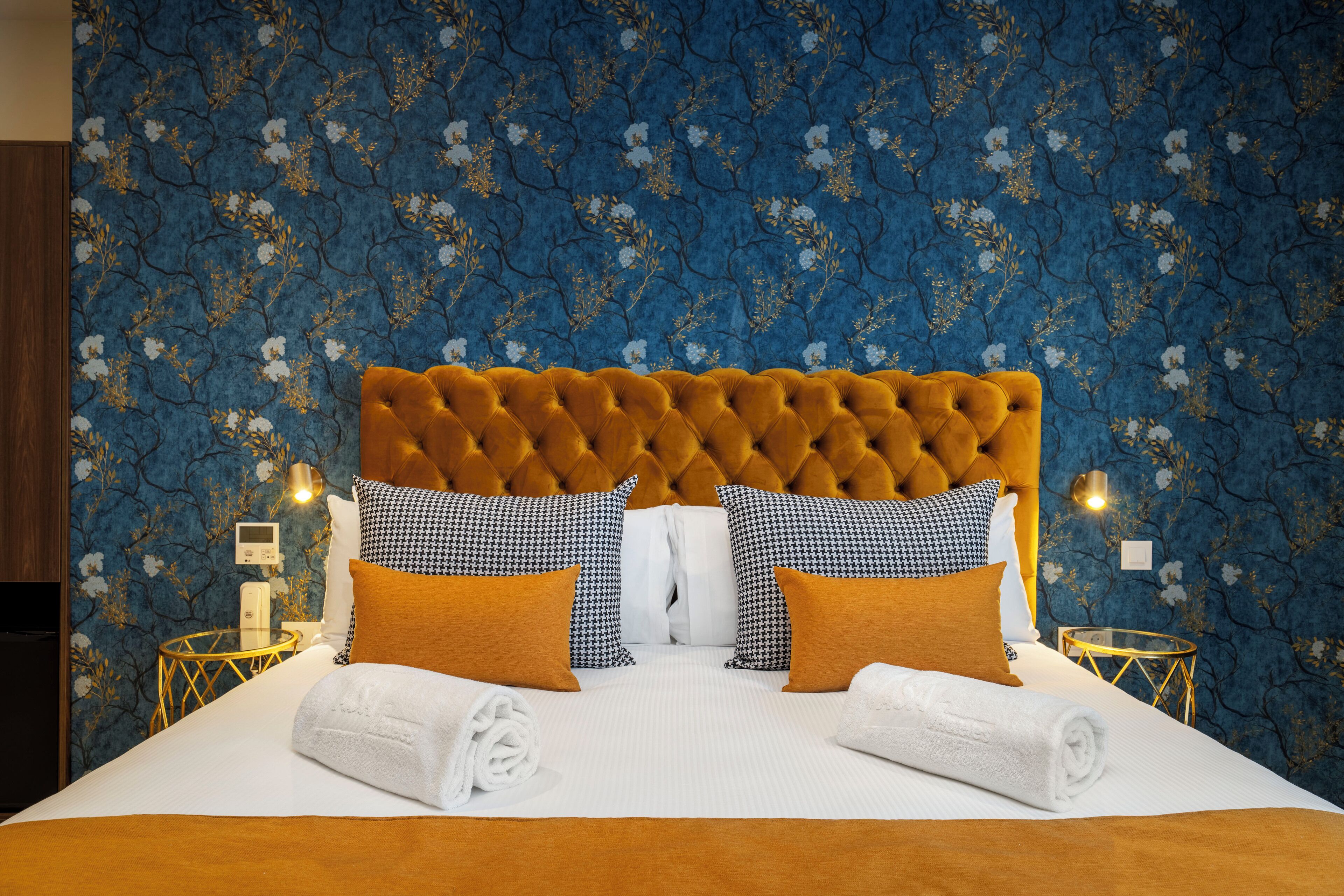 A hotel room with a tufted headboard, a large bed with white linens, two gold bedside tables, and a blue floral wallpaper.
