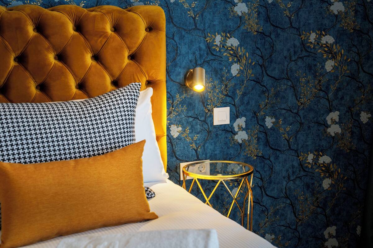Une chambre à coucher avec une tête de lit capitonnée, un coussin à motif pied-de-poule, une table de chevet avec un cadre doré et un mur orné d’un motif floral.