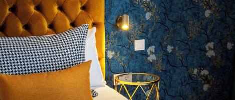 Un dormitorio con cabecera capitonada, almohada de pata de gallo, mesita de noche con marco dorado y pared con estampado floral.