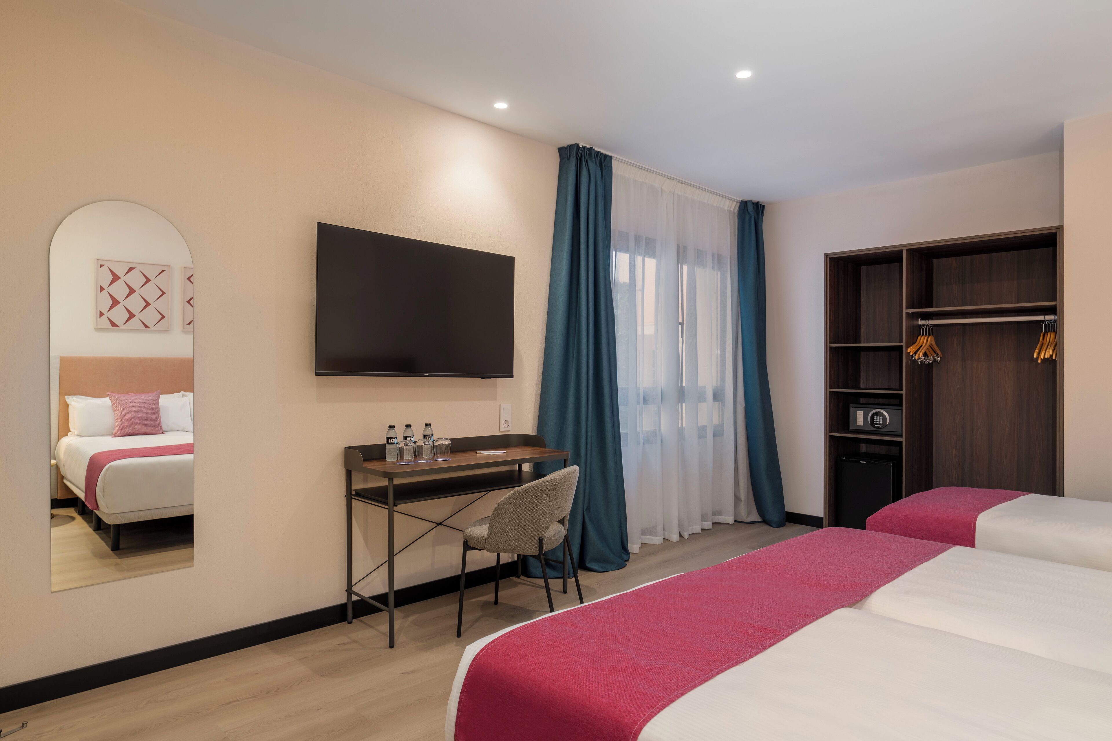 A hotel room with two beds, a desk with a chair, a TV mounted on the wall, a wardrobe, and a mirror reflecting the bed.