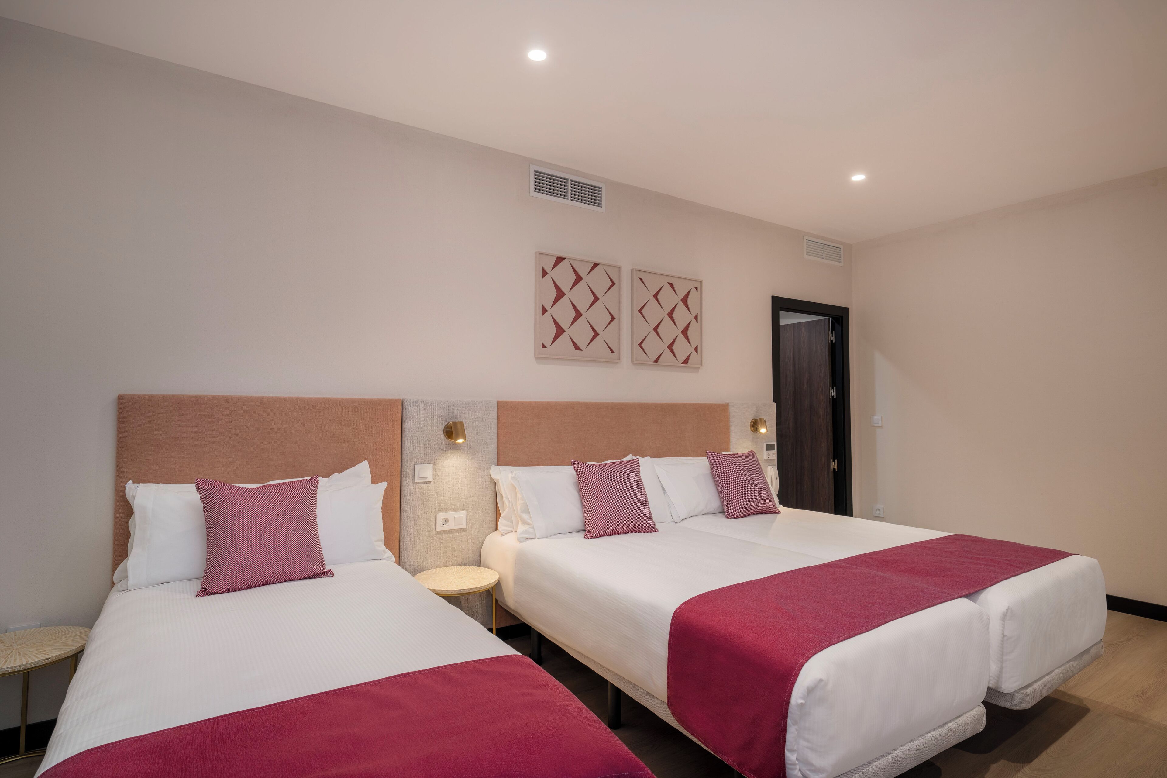 Habitación de hotel con dos camas, cada una con ropa blanca y mantas rojas. Hay lámparas fijadas a la pared y una puerta con marco oscuro.