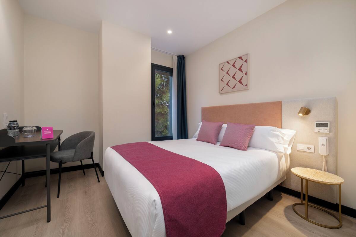 Une chambre d’hôtel comprenant un lit, un bureau avec une chaise, une table de chevet, une fenêtre avec des rideaux et un thermostat fixé au mur.